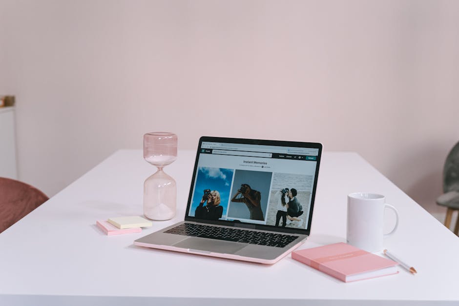 A clean and minimalist workspace featuring a laptop, hourglass, notebook, and coffee mug on a white desk.