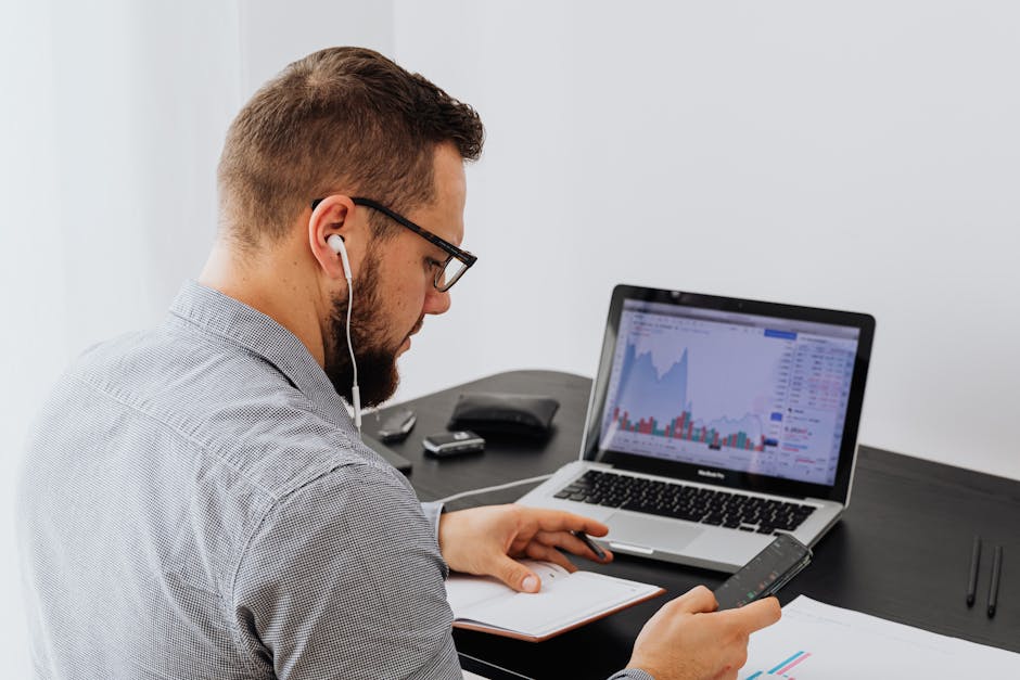A businessman with earphones analyzes stock market graphs on a laptop while using a smartphone and notebook.