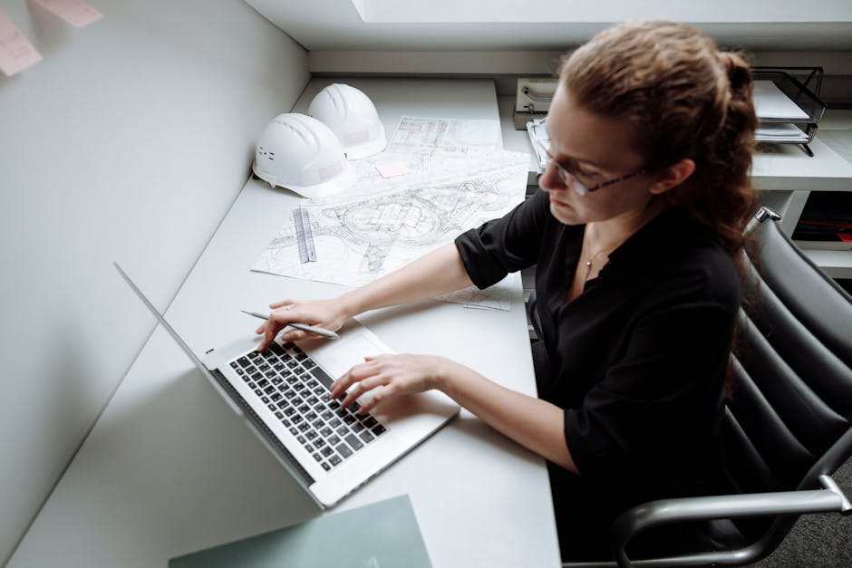 Woman architect working on a laptop at her desk with blueprints and safety helmets.
