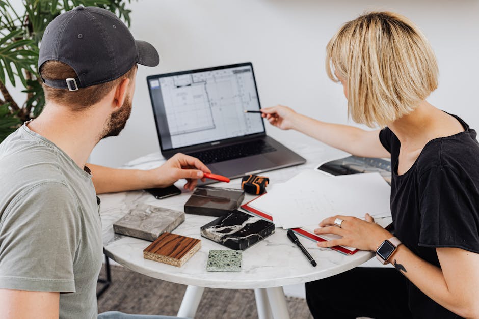 Two designers discussing building plans with laptop and samples in office.