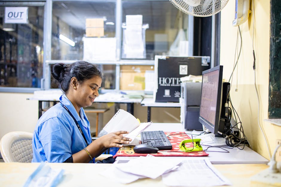 A woman in an office setting reading paperwork at her desk with a computer.